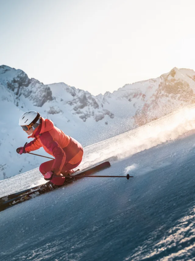 Alpine skier descending a slope with a panoramic view of the Aravis mountain range in the background. The sky is fairly clear, with a lovely golden glow moving behind the skier, wearing a pink ski suit.