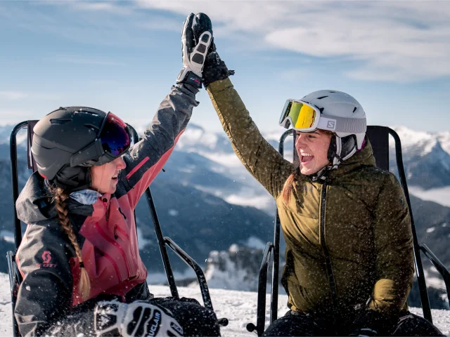 2 friends in skiwear are sitting on deckchairs at the top of a snow-covered mountain, smiling as they high-five each other. There's a panoramic view of the Aravis mountain range in the background. They are enjoying a moment of relaxation under a sunny sky. One person is wearing a pink jacket, another a green one. The atmosphere is friendly and relaxed, reminiscent of a break after a skiing session.