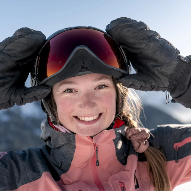 Close-up of a smiling young woman wearing a pink and grey ski jacket, black gloves and a helmet with ski goggles. She is adjusting her glasses with her hands. In the background are snow-capped mountains under a blue sky. Her radiant expression and the alpine setting give an impression of joy and pleasure in the mountains.