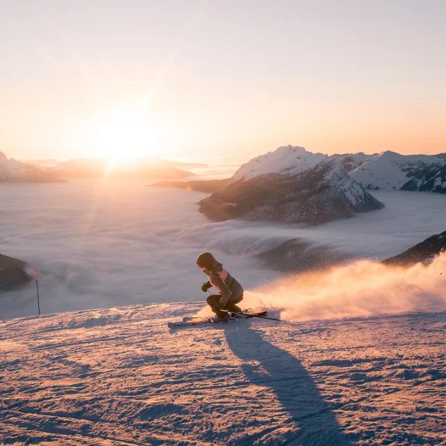 Une skieuse dévale une pente enneigée au coucher du soleil, projetant un nuage de poudreuse derrière elle. En arrière-plan, une mer de nuages enveloppe les vallées, tandis que le mont Bagy se découpe dans une lumière dorée, créant une scène spectaculaire et apaisante.