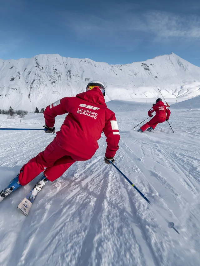 Zwei ESF-Skilehrer fahren eine Skipiste ziemlich langsam hinunter. Sie sind mit einem roten Skianzug in den Farben der ESF bekleidet. Wir sehen die Aravis-Kette und einen blauen, leicht bewölkten Himmel im Hintergrund. Die Atmosphäre ist dynamisch und sanft zugleich.