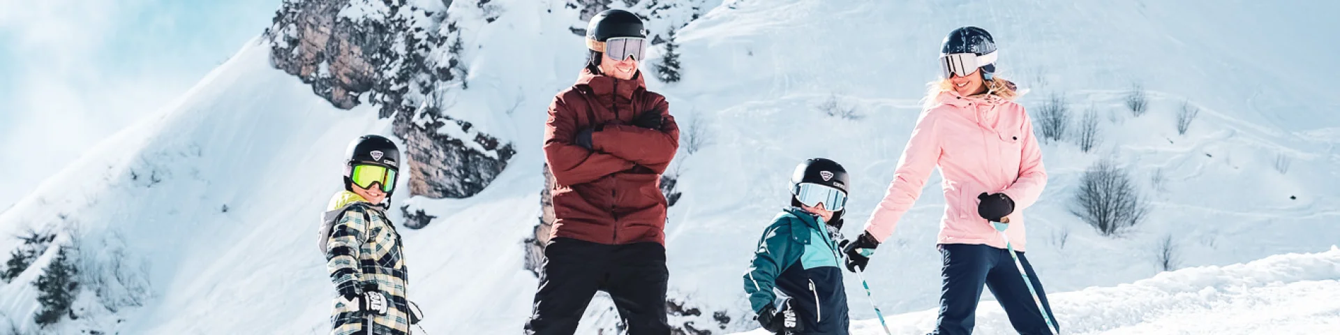 A family consisting of a father in red and black ski clothes and a snowboard, a mother in pink and black ski clothes and two little boys in multicoloured ski clothes, pose on skis in front of the Pointe Percée with big smiles on their faces. They're ready to hit the slopes. The image has a lovely atmosphere, with white snow and a slightly cloudy blue sky.