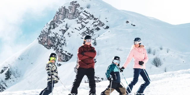 A family consisting of a father in red and black ski clothes and a snowboard, a mother in pink and black ski clothes and two little boys in multicoloured ski clothes, pose on skis in front of the Pointe Percée with big smiles on their faces. They're ready to hit the slopes. The image has a lovely atmosphere, with white snow and a slightly cloudy blue sky.