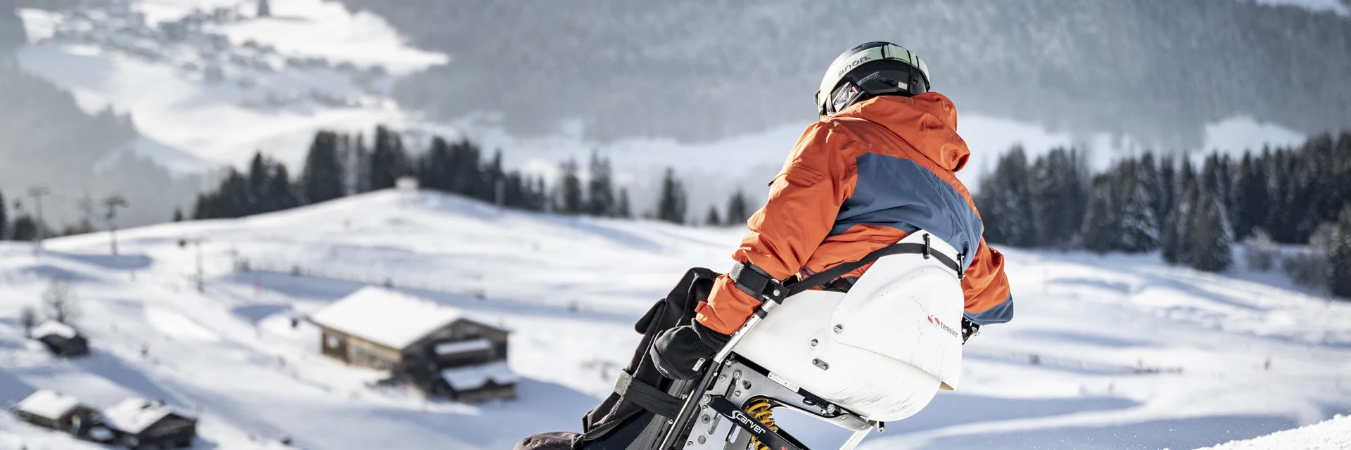 A person with reduced mobility goes handiskiing in a snowy setting. The skier is wearing a black, orange and grey ski suit with a black helmet. We can see the Bargy massif in the background. The atmosphere is soft and serene.