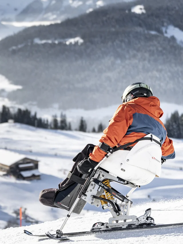A person with reduced mobility goes handiskiing in a snowy setting. The skier is wearing a black, orange and grey ski suit with a black helmet. We can see the Bargy massif in the background. The atmosphere is soft and serene.