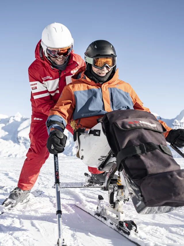 Une personne à mobilité réduite pratique le handiski dans un décor enneigé. Son accompagnateur de l'ESF en habits rouges le pousse et l'assure derrière lui, tous deux rigolent et semblent apprécier ce moment. Derrière eux, la femme du skieur PMR les suit. Le skieur PMR est vêtu d'un ensemble de ski noir, orange et gris avec un casque noir. Nous pouvons apercevoir la chaîne des Aravis en arrière-plan. L'atmosphère est très douce, serène et joviale.