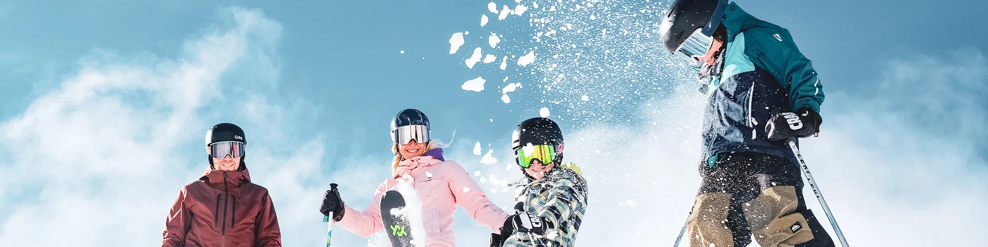 A family of four in colourful ski outfits and ski equipment including gloves, helmets and goggles, in a snowy environment under a clear blue sky. The people are positioned at the top of a snow-covered slope. A child in the centre throws snow into the air with his ski, creating a lively and joyful dynamic.
