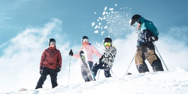 Une famille de quatre personnes en tenue de ski colorées ainsi que des équipements de ski comprenant des gants, des casques et des lunettes de protection, dans un environnement enneigé sous un ciel bleu clair. Les personnes sont placées en haut d'une pente enneigée. Un enfant au centre projette de la neige en l'air avec son ski, créant une dynamique joyeuse et animée.