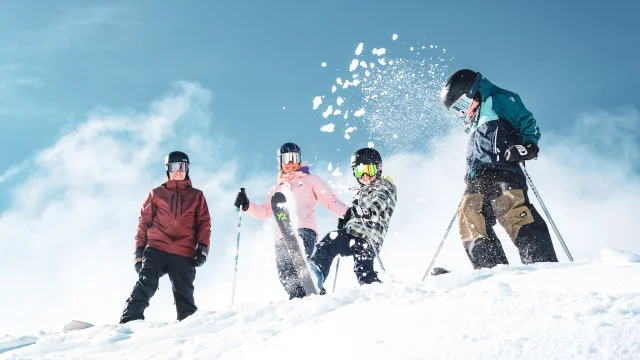 Une famille de quatre personnes en tenue de ski colorées ainsi que des équipements de ski comprenant des gants, des casques et des lunettes de protection, dans un environnement enneigé sous un ciel bleu clair. Les personnes sont placées en haut d'une pente enneigée. Un enfant au centre projette de la neige en l'air avec son ski, créant une dynamique joyeuse et animée.