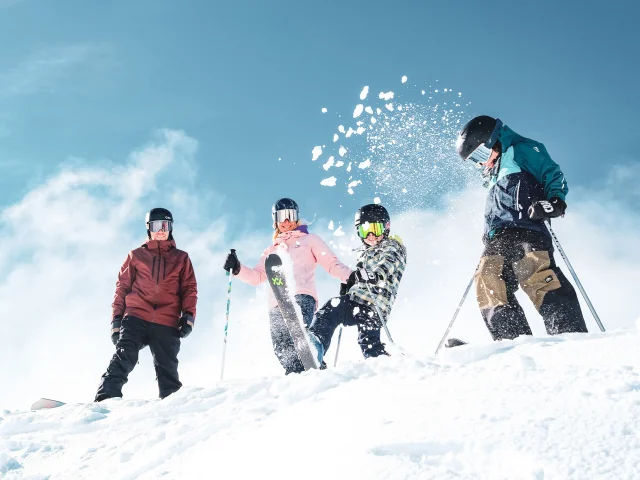 A family of four in colourful ski outfits and ski equipment including gloves, helmets and goggles, in a snowy environment under a clear blue sky. The people are positioned at the top of a snow-covered slope. A child in the centre throws snow into the air with his ski, creating a lively and joyful dynamic.
