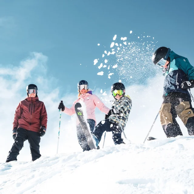 A family of four in colourful ski outfits and ski equipment including gloves, helmets and goggles, in a snowy environment under a clear blue sky. The people are positioned at the top of a snow-covered slope. A child in the centre throws snow into the air with his ski, creating a lively and joyful dynamic.
