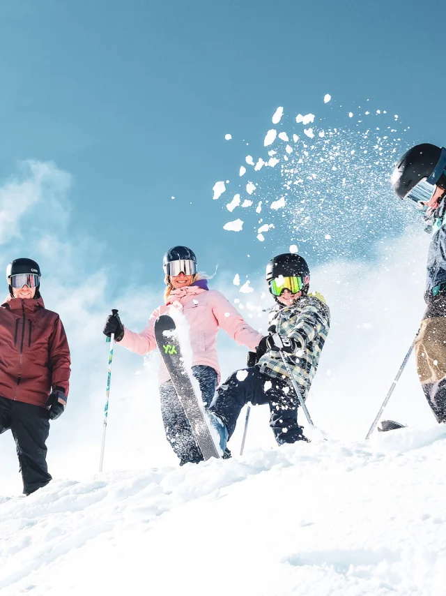 A family of four in colourful ski outfits and ski equipment including gloves, helmets and goggles, in a snowy environment under a clear blue sky. The people are positioned at the top of a snow-covered slope. A child in the centre throws snow into the air with his ski, creating a lively and joyful dynamic.