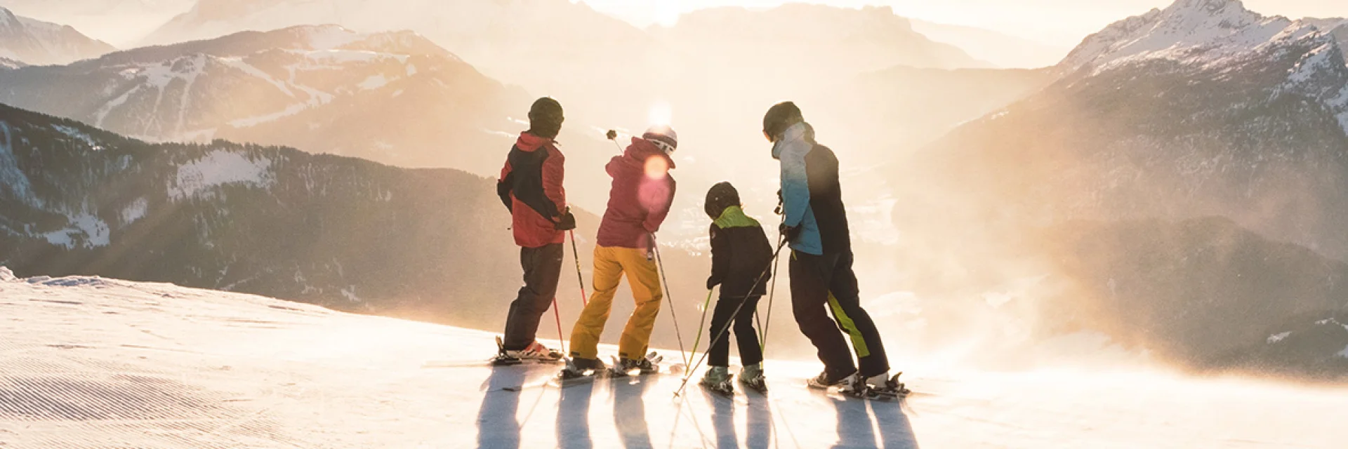 Une famille composée de deux enfants se tenant sur des skis en haut d'une piste contemple le coucher de soleil avec vue sur la chaîne des Aravis. L'un a un ensemble de ski bleu, noir et vert, un autre rouge et jaune, un autre vert et noir et le dernier rouge et noir. L'un des skieurs au milieu pointe avec son bâton gauche le sommet d'une montagne. L'atmosphère est très lunaire avec une lumière blanche/dorée. C'est une belle fin de journée de ski.