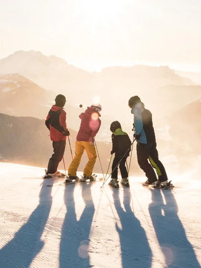 A family with two children standing on skis at the top of a slope watching the sun set over the Aravis mountain range. One has a blue, black and green ski outfit, another red and yellow, another green and black and the last red and black. One of the skiers in the middle is pointing with his left stick to the top of a mountain. The atmosphere is very lunar with a white/golden light. It's a beautiful end to a day's skiing.