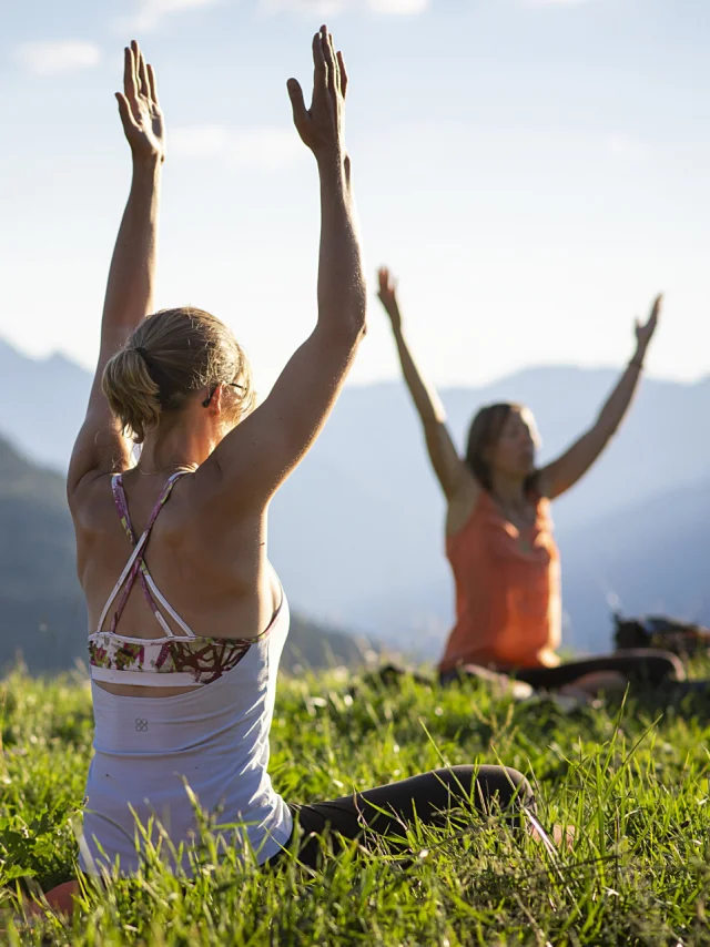 A group of 3 people sitting on the grass raise their hands to the sky in a yoga position, facing a mountainous landscape under a blue sky.