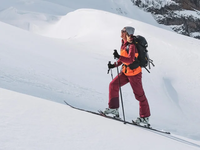 Une skieuse souriante en randonnée remonte la pente sous les sommets enneigés