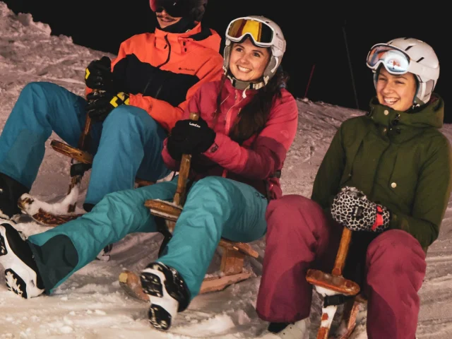 3 people dressed warmly are each sitting on a tray with a smile, the photo is taken at night.