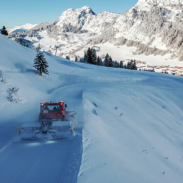 Une dameuse rouge travaille sur une piste enneigée sinueuse, entourée de montagnes enneigées et de sapins, avec le village du chinaillon visible en contrebas.