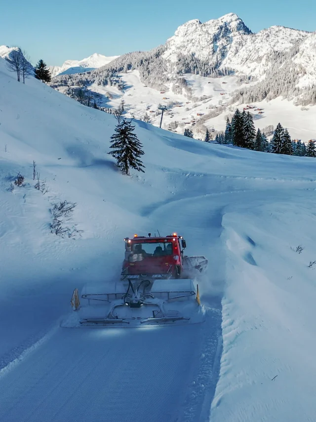A red groomer works its way down a winding snowy slope, surrounded by snow-capped mountains and fir trees, with the village of Chinaillon visible below.