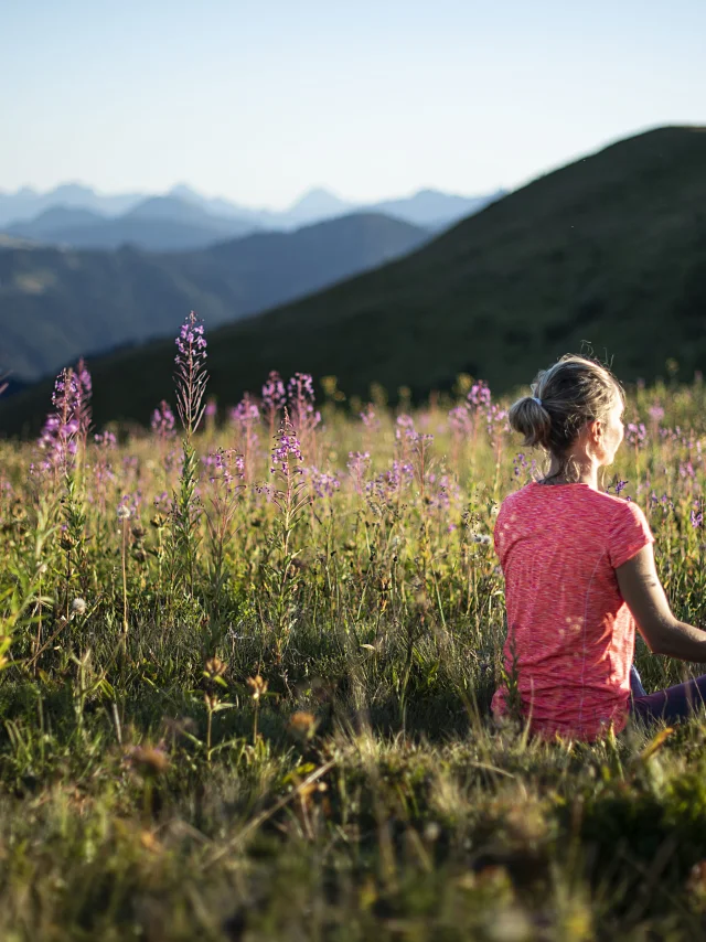 A woman practising yoga in a green mountain field, in the lotus posture, surrounded by violet flowers.