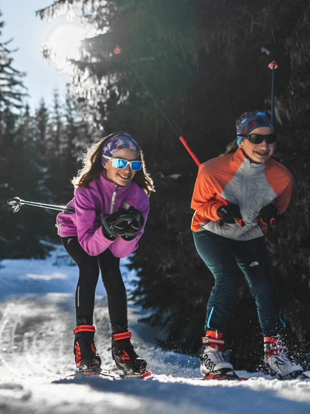 Three smiling, well-equipped cross-country skiers glide down a snow-covered track through a fir forest in the winter sunshine.