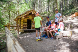 groupe d'enfants assis sur un banc au milieu de la foret à côté d'une cabane en rondin